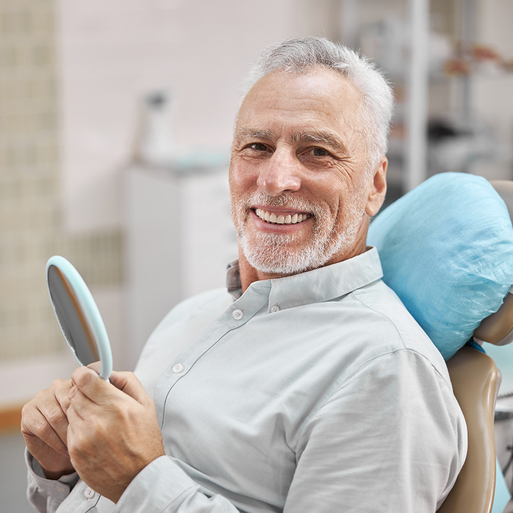 The image shows a man sitting in a dental chair with a large mirror in front of him, smiling at the camera, wearing a beard and mustache, and appears to be in a dental office setting.