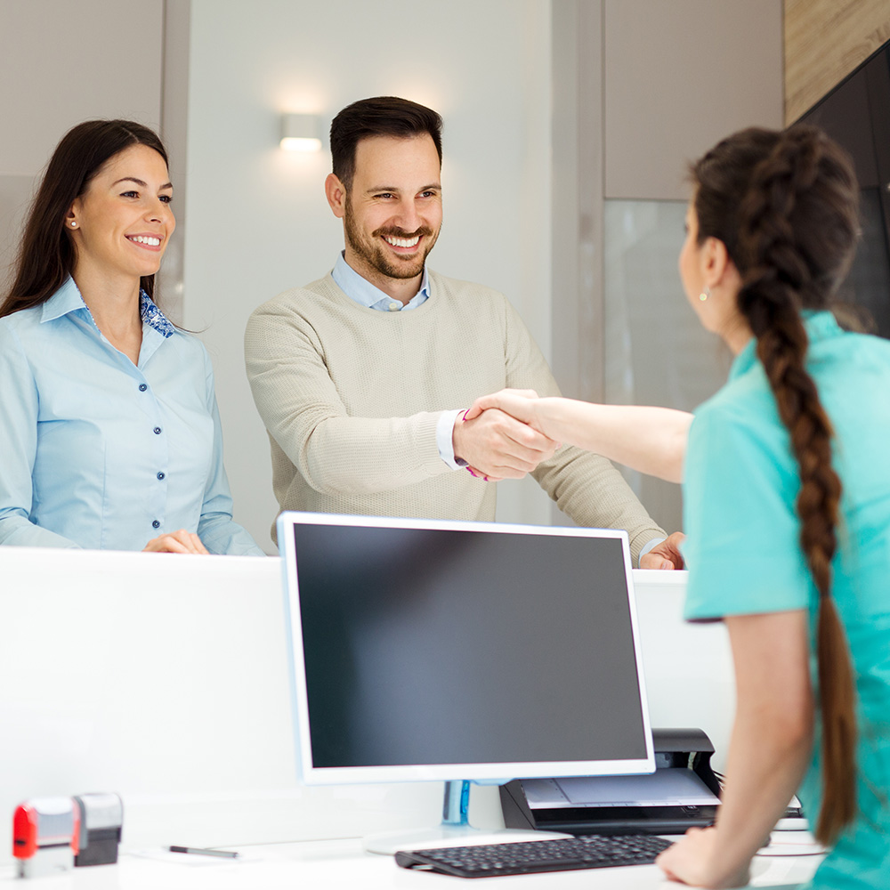 The image shows three individuals in a professional setting, with two adults shaking hands over a desk while another person appears to be seated at the desk, possibly receiving assistance or service from the shakers of hands.