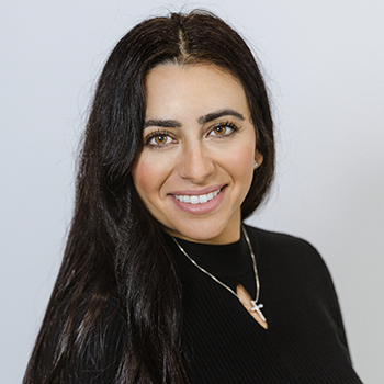 The image shows a woman with long hair, wearing a black top and a necklace, posing for a portrait with a smile.